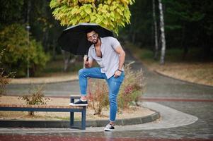 hombre de barba árabe alto de moda usa camisa, jeans y gafas de sol con paraguas posado bajo la lluvia en la plaza del parque. foto