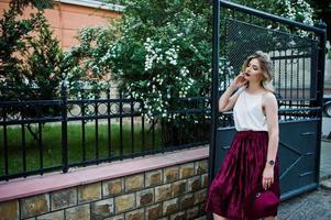 Fashionable and beautiful blonde model girl in stylish red velvet velour skirt, white blouse and  hat, posing outdoor against gates. photo