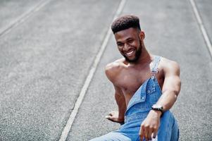 Handsome sexy african american bare torso man at jeans overalls sitting at stadium racecourse. Fashionable black man portrait. photo