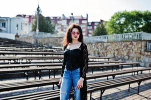 retrato de una chica morena con gafas rosas puestas en negro al aire libre en un día soleado contra una fila de bancos. foto