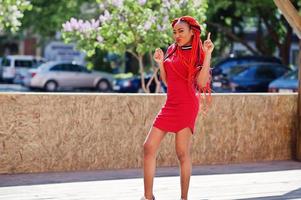 Cute and slim african american girl in red dress with dreadlocks in motion having fun on street. Stylish black model. photo