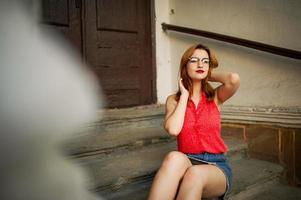 atractiva mujer pelirroja con anteojos, vestida con blusa roja y falda de jeans posando. foto