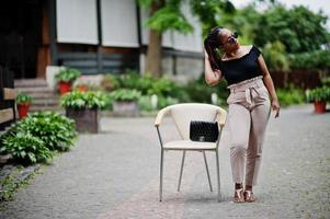 Stylish african american woman in sunglasses posed outdoor at chair. photo