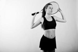 Black and white portrait of beautiful young woman player in sports clothes holding tennis racket while standing against white background. photo