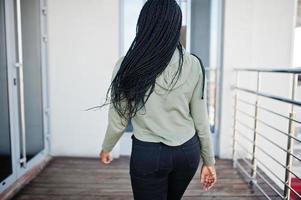 City portrait of positive young dark skinned female wearing green hoody and eyeglasses standing at balkony. photo