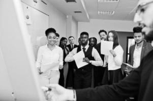 Arabian business coach presenting report standing near whiteboard pointing on sales statistic shown on diagram and chart teach diverse multiracial company members gathered together in conference room. photo