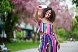 Fashionable african american woman in pink striped jumpsuit posed at spring bloom street and making selfie by mobile phone.