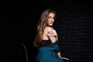 Handsome brunette girl wear on black and green jacket, sitting and posing on chair at studio against dark brick wall. Studio model portrait. photo