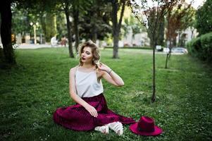 Fashionable and beautiful blonde model girl in stylish red velvet velour skirt, white blouse and hat, sitting on green grass at park. photo