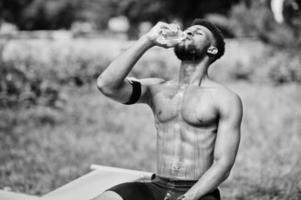 African american male athlete sexy sport bare torso man with running sports arm case for mobile phone, doing exercises on the street workout simulators and drinking water from bottle. photo