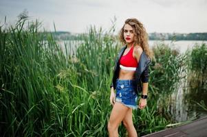 Sexy curly model girl in red top, jeans denim shorts, leather jacket and sneakers posed in the reed. photo