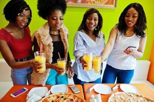 Four young african girls in bright colored pizza restaurant clinking juices. photo