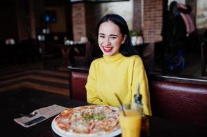 chica morena divertida en suéter amarillo comiendo pizza en el restaurante. foto