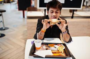 hombre indio con estilo sentado en un café de comida rápida y comiendo hamburguesas. mostrar su lengua. foto