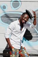 Stylish african american man in white shirt and colored pants with hat and glasses posed outdoor against graffiti wall. Black fashionable model boy. photo
