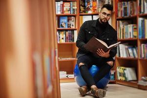 Tall smart arab student man, wear on black jeans jacket and eyeglasses, sitting at library and read book at hands. photo