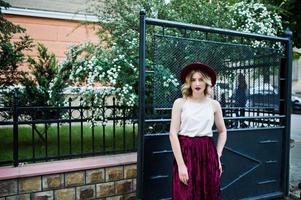 Fashionable and beautiful blonde model girl in stylish red velvet velour skirt, white blouse and  hat, posing outdoor against gates. photo