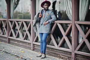 Stylish African American man model in gray coat, jacket tie and red hat posed against wooden cafe. photo