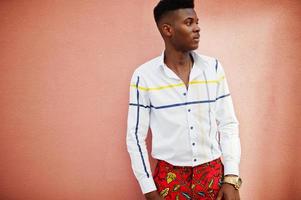 Portrait of handsome stylish african american model man in red throusers and white shirt against pink wall. photo