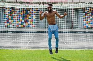 Handsome sexy african american muscular bare torso man at jeans overalls posed at green grass agasinst football gates of stadium field. Fashionable black man portrait. photo