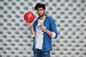 Stylish asian man in jeans shirt standing with bowling ball at hand against silver wall background and show two fingers. photo