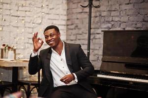 Strong powerful african american man in black suit sitting against piano and show ok sign. photo