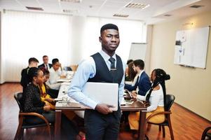 Face of handsome african business man, holding laptop on the background of business peoples multiracial team meeting, sitting in office table. photo