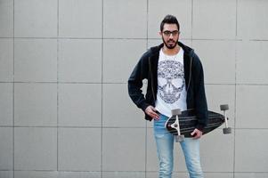 Street style arab man in eyeglasses with longboard posed against gray wall. photo