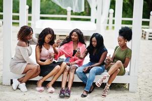 Group of five african american girls relaxing at beautiful poolside cabana beside luxury resort and looking at mobile phones. photo