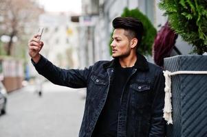 Handsome and fashionable indian man in black jeans jacket posed outdoor and making selfie on phone. photo