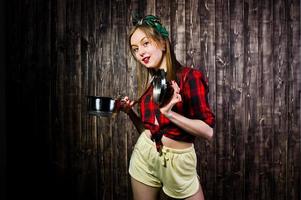 Young funny housewife in checkered shirt and yellow shorts pin up style with saucepan on wooden background. photo