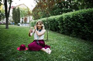 chica modelo rubia de moda y hermosa con elegante falda de terciopelo rojo, blusa blanca y sombrero, sentada en la hierba verde en el parque con teléfono y auriculares. ella hace autofoto. foto