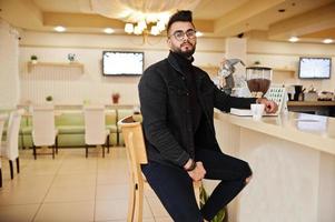 el hombre árabe usa una chaqueta negra de jeans y anteojos en un café bebe café en el bar con un libro. chico modelo árabe elegante y de moda. foto