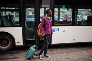 African american man in checkered shirt, with suitcase and backpack. Black man traveler against bus. photo