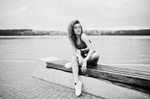 Sexy curly model girl in red top, jeans denim shorts, eyeglasses and sneakers posed on bench against lake. photo