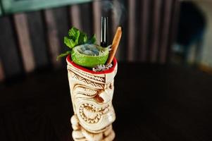 Alcoholic cocktail with mint and lime with fire in originally voodoo earthen glass on bar table. photo