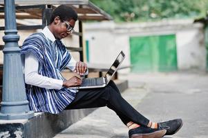 African man in traditional clothes and glasses with laptop working outdoor and looking at time of his watches. photo