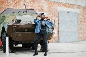 African american man in jeans jacket, beret and eyeglasses, with cigar posed against btr military armored vehicle, and making selfie on mobile phone. photo