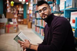Tall smart arab student man, wear on violet turtleneck and eyeglasses, at library sitting against books shelves. photo
