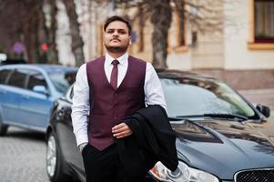 Elegante hombre de negocios indio con traje formal de chaleco de pie contra un coche de negocios negro en la calle de la ciudad. foto