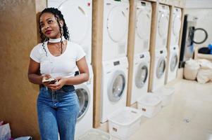 alegre mujer afroamericana cerca de la lavadora escuchando música con auriculares desde el teléfono móvil en la lavandería de autoservicio. foto