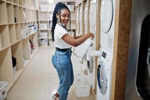 alegre mujer afroamericana sostiene detergente cerca de la lavadora en la lavandería de autoservicio. foto