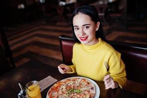 chica morena divertida en suéter amarillo comiendo pizza en el restaurante. foto