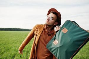 hombre elegante con gafas, chaqueta marrón y sombrero con bolso posado en campo verde. foto