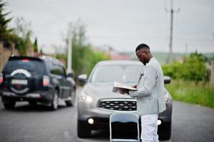 hombre afroamericano rico y elegante en blazer y pantalones blancos, anteojos lee la revista contra su auto todoterreno. foto