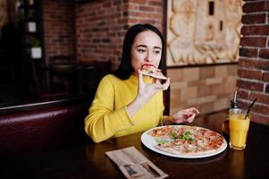 Funny brunette girl in yellow sweater eating pizza at restaurant. photo