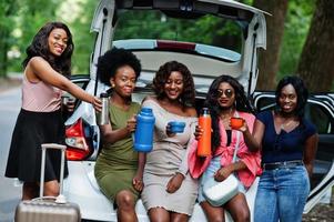 Group of five happy african american traveler girls sitting in car open trunk and drink tea from thermos. photo