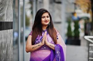 Indian hindu girl at traditional violet saree posed at street and shows namaste tatooed hands sign. photo