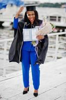 chica estudiante afroamericana en vestido de graduación negro con diploma, en traje azul posado. foto
