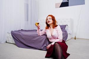 Gorgeous red haired girl in pink blouse and red skirt with glass of wine at hand near bed at room. photo
